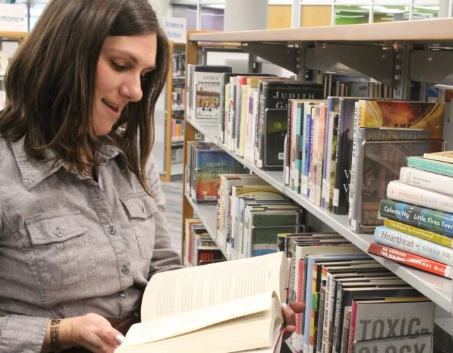 A person is standing next to a book shelf and looking at an open book. A short stack of books is on the shelf.