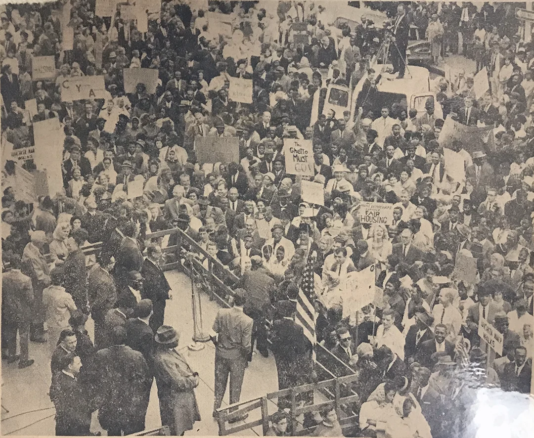 A photograph of a crowd in front of a stage. Some people are holding signs.