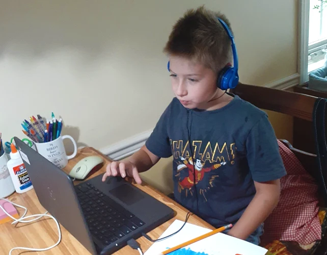 A student attending remote school during the 2020-21 school year is sitting at a desk inside a home while wearing headphones and looking at the screen on his school-issued Chromebook