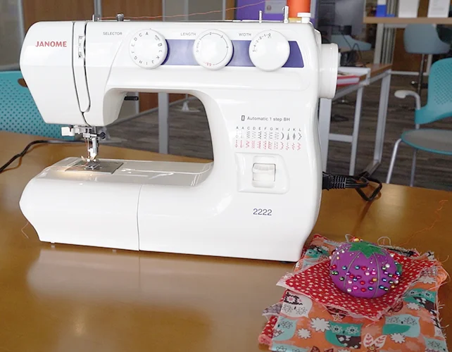 The Janome 2222 sewing machine on a table in the Advanced Learning Library. To the right of the machine is a small stack of scrap fabric underneath a pincushion with numerous pins sticking out.