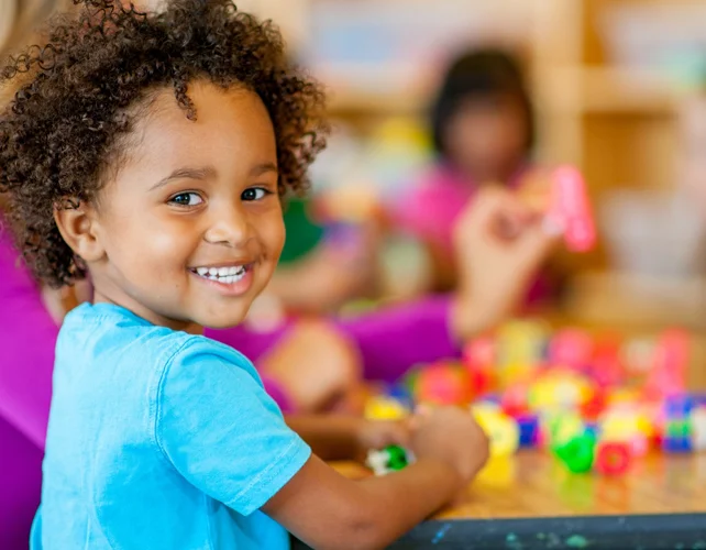 A child is at a table with several small pieces of a toy that might be building blocks. The child is looking over their shoulder at the camera.